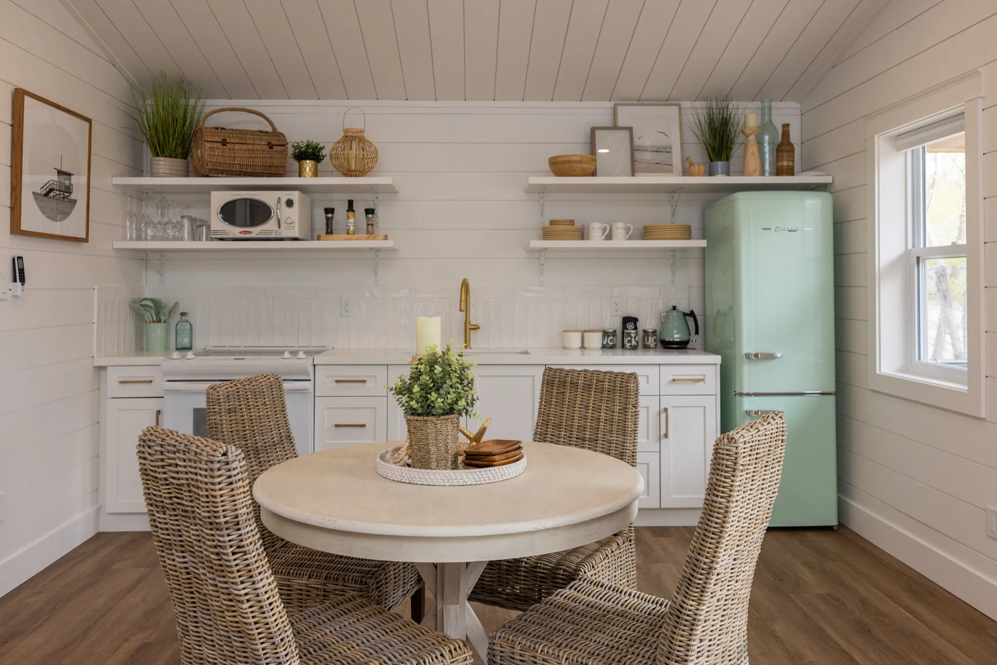 The Beach cabin's kitchen and dining area, with white shiplap walls, open shelving, a mint-green retro fridge and a round table with woven wicker chairs.
