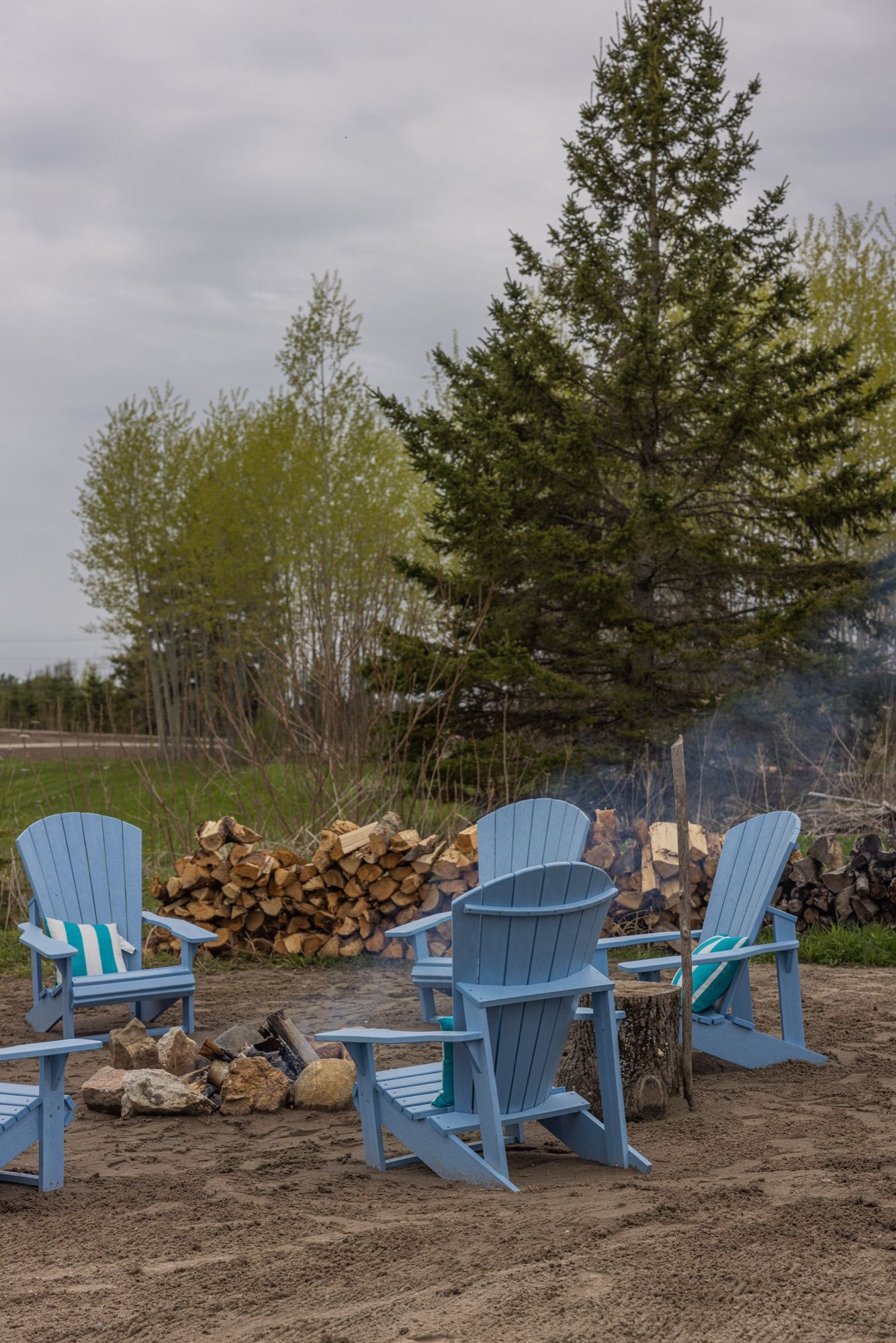 Blue Adirondack chairs set around a stone fire ring on the sand, with a stacked woodpile and evergreens behind.